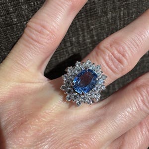 May include: A large, oval-cut, light blue gemstone ring with a halo of smaller, clear gemstones. The ring is set in a silver-colored metal and is displayed on a person's finger. The ring's design is classic and elegant.