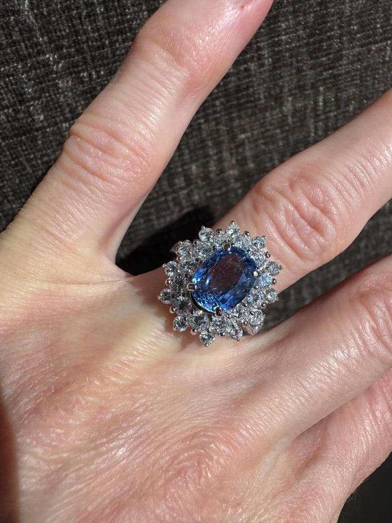 May include: A large, oval-cut, light blue gemstone ring with a halo of smaller, clear gemstones. The ring is set in a silver-colored metal and is displayed on a person's finger. The ring's design is classic and elegant.