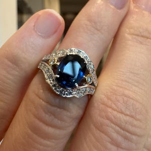 May include: A silver ring with a large, oval, deep blue gemstone as the centerpiece. The ring is surrounded by smaller, clear gemstones and gold accents. The design is intricate and elegant, suitable for special occasions.
