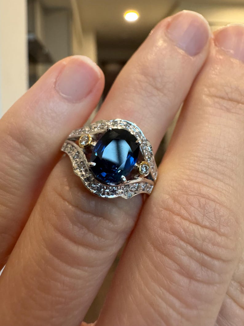 May include: A silver ring with a large, oval, deep blue gemstone as the centerpiece. The ring is surrounded by smaller, clear gemstones and gold accents. The design is intricate and elegant, suitable for special occasions.
