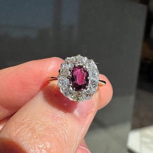 May include: A gold ring featuring a central oval-cut ruby surrounded by a halo of white and yellow gemstones. The ring's design is classic and elegant, with a focus on the vibrant red of the ruby.