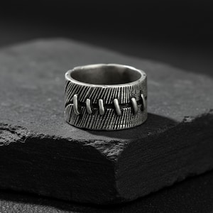 May include: A silver-toned ring with a textured surface and a stitched design. The ring has a wide band with vertical lines and a series of horizontal stitches across the front. The ring is displayed on a dark, textured surface.
