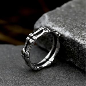 Könnte beinhalten: Ein silberfarbener Ring mit einem Skelettknochen-Design. Der Ring zeigt eine detaillierte Darstellung von Knochen und erzeugt eine einzigartige und gotische Ästhetik. Der Ring wird auf einer grauen Oberfläche gezeigt.