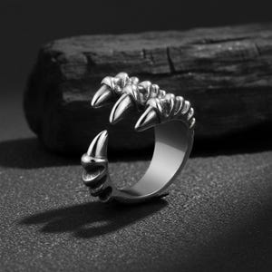 Könnte beinhalten: Ein silberfarbener Krallenring mit scharfen, spitzen Spitzen. Der Ring hat ein detailliertes Design einer Klaue mit strukturierten Rippen und einer polierten Oberfläche. Die Klaue umschließt den Finger und schafft ein auffälliges Accessoire.