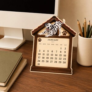 May include: A wooden desk calendar shaped like a doghouse with a dalmatian puppy illustration on top. The calendar displays the month of January 2027 with black text on a light tan background, featuring paw print accents and a dark brown wood frame.