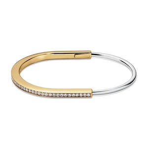 Puede incluir: Una pulsera bicolor dorada y plateada. La parte dorada está incrustada con pequeñas piedras preciosas transparentes. La pulsera tiene un cierre de bisagra. El diseño es de estilo moderno y minimalista.