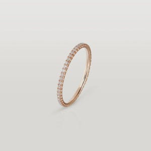 Puede incluir: Un anillo de eternidad de oro rosa con una fila continua de peque&ntilde;as piedras preciosas claras. El anillo est&aacute; sobre un fondo suave y neutro, resaltando su dise&ntilde;o delicado y brillo. La banda es delgada y elegante.