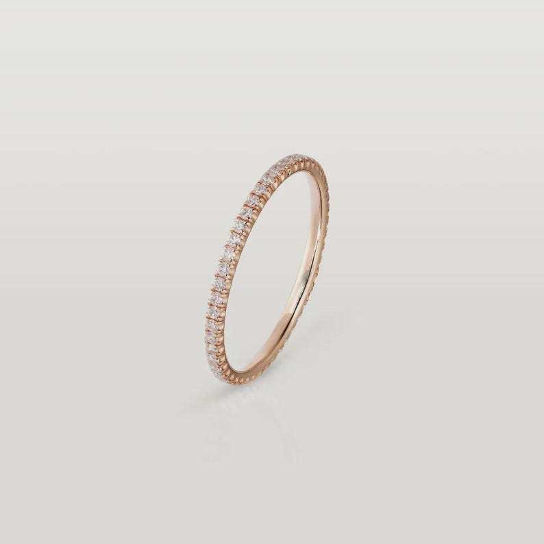 Puede incluir: Un anillo de eternidad de oro rosa con una fila continua de peque&ntilde;as piedras preciosas claras. El anillo est&aacute; sobre un fondo suave y neutro, resaltando su dise&ntilde;o delicado y brillo. La banda es delgada y elegante.