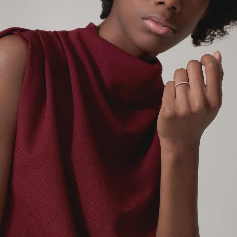 Puede incluir: Un delicado anillo con peque&ntilde;as piedras brillantes se lleva en un dedo. El anillo se destaca sobre un fondo de una blusa sin mangas de color rojo intenso. La imagen es un primer plano, que resalta los detalles del anillo y el rico color de la tela.