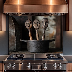 Op de afbeelding: Drie houten lepels met gebeeldhouwde gezichten staan in een zwarte gietijzeren pot. De lepels staan voor een roestvrijstalen fornuis en afzuigkap. De keuken heeft houten kasten en een donker aanrecht.