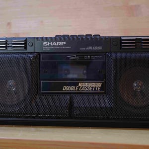 Puede incluir: Grabadora de casete de radio estéreo Sharp negra. Cuenta con una pletina de doble casete, ecualizador gráfico de 3 bandas y dos altavoces. El texto "Twin Mechanism Double Cassette" es visible.