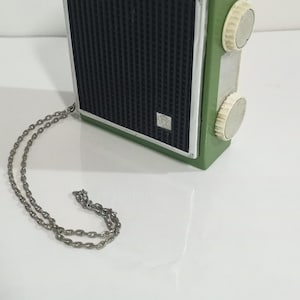 Puede incluir: Una radio portátil con carcasa rectangular de plástico verde, rejilla de altavoz negra y marco metálico plateado. Cuenta con dos perillas de control circulares blancas en el lateral y una fina cadena de metal plateado unida a la esquina.