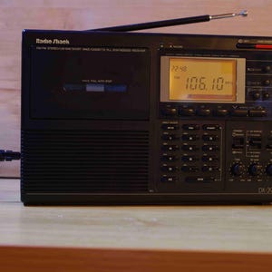 May include: Black Radio Shack DX-322 receiver with an extendable aerial. The radio has a digital display showing the time and radio frequency. The radio has a cassette player and is plugged into a power source.