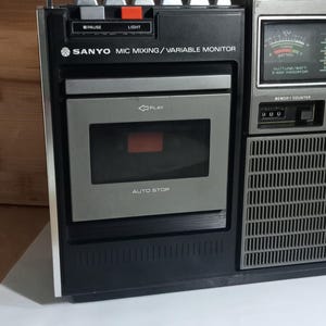 Op de afbeelding: Een zwarte en zilverkleurige draagbare Sanyo-cassettespeler met een handvat aan de bovenkant, een centraal cassettedeck met auto-stopfunctie, een VU-meter en zilveren bedieningsknoppen. Het apparaat bevat tekstlabels voor mic mixing en variable monitor.