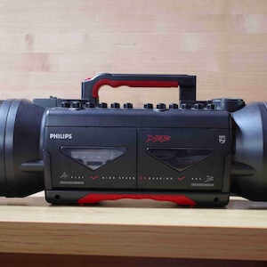 May include: Black Philips boombox with red detailing, featuring large speakers. The boombox has a handle and control buttons on top. The text "Philips" and "D83" are visible on the front.