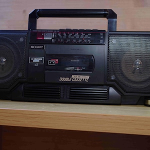 Puede incluir: Boombox negro Sharp con altavoces grandes, un asa y un reproductor de casetes. La radio tiene un dial de sintonización y las palabras "X-BASS" y "DOUBLE CASSETTE" son visibles.