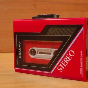 Puede incluir: Un reproductor de casetes estéreo Sanyo rojo. El reproductor tiene un diseño negro y dorado alrededor de la ventana del casete y la palabra "STEREO" en blanco. El reproductor de casetes está sobre una superficie de madera.