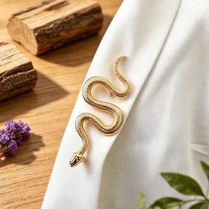 May include: A gold-toned metal brooch shaped like a coiled snake with a textured, scale-like surface. The snake is depicted in an S-shaped curve, featuring a detailed head and a slender, tapering tail, pinned securely onto a smooth white fabric.