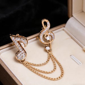 Puede incluir: Broche dorado con forma de nota musical y clave de sol con detalles de cristal transparente. El broche tiene una doble cadena y se muestra en una caja de joyería beige. Un accesorio elegante para los amantes de la música.
