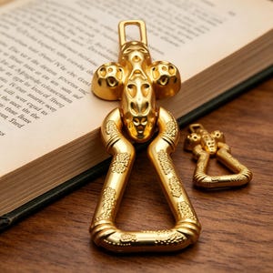 Peut inclure: Deux porte-clés dorés aux motifs complexes. Le plus grand a une partie supérieure en forme de croix et une base triangulaire, tandis que le plus petit reprend le design. Ils sont posés sur une surface en bois, à côté d'un livre ouvert.