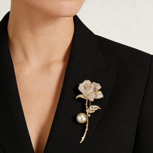 May include: A gold-coloured rose brooch with sparkling crystal petals and a pearl accent. The stem and leaves are also gold-coloured. The brooch is pinned to a black blazer.