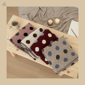 May include: A collection of folded blankets with large polka dots in various colors, including pink, cream, burgundy, and gray, arranged on a wooden surface. Each blanket features a different color of polka dots. A cup of coffee, candles, and a notepad are also visible.