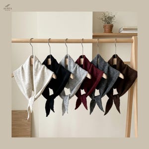 May include: A display of seven triangular knit scarves in various colors, including beige, black, gray, burgundy, and brown, hanging on a wooden rack. Each scarf has a tie closure. The brand name "AERIA STUDIO" is visible in the top left corner.