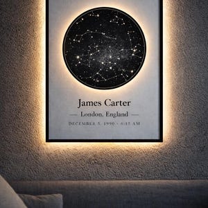 May include: Framed star map with a black background and white constellations. The frame is black with a warm, glowing light around it. The text on the map reads "James Carter, London, England, December 5, 1990, 6:15 AM."