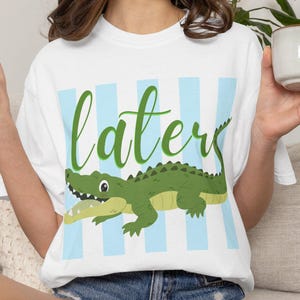 Peut inclure: T-shirt blanc avec un motif d'alligator vert et le mot "later" en écriture verte. Le t-shirt présente des rayures verticales bleu clair. Le modèle porte un short en jean.