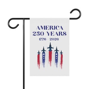 Peut inclure: Un drapeau de jardin blanc avec le texte "AMERICA 250 YEARS 1776-2026" en bleu marine. En dessous, cinq avions stylisés avec des traînées de vapeur rouges, blanches et bleues. Le drapeau est monté sur un support en métal noir.