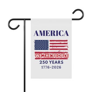 Peut inclure: Un drapeau de jardin blanc avec le mot "AMERICA" en bleu, le drapeau américain et le texte "WE THE PEOPLE 250 YEARS 1776-2026" en rouge. Le drapeau est monté sur un support en métal noir.