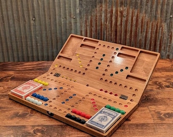 May include: A wooden cribbage board game, open to display the playing surface. The board has holes for pegs, with playing cards and pegs in various colors. The game is set on a wooden surface.