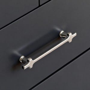 May include: A dark grey cabinet with silver-coloured handles and knobs. The cabinet has a long, silver handle and two smaller cross-shaped knobs. A cream-coloured, textured fabric is partially visible.