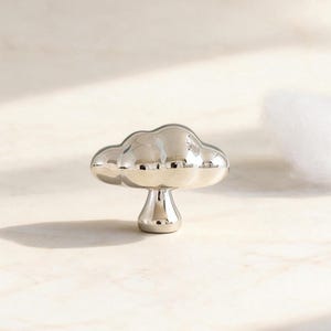 May include: A polished silver cloud-shaped cabinet knob. The knob is positioned centrally on a light surface, with two fluffy white cotton clouds on either side. The knob has a rounded, three-dimensional design.