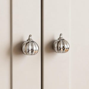 Könnte beinhalten: Zwei silberne, kürbisförmige Schrankknöpfe an einer cremefarbenen Schranktür. Die Knöpfe haben eine gerippte Textur und einen kleinen Stiel. Die Schranktüren sind schlicht und haben ein vertikales Paneeldesign.