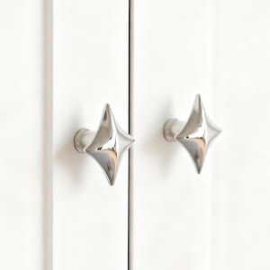 May include: Two polished chrome star-shaped cabinet knobs mounted on a white cabinet door. The knobs have a four-pointed star design with a reflective surface, adding a modern touch to the cabinetry.