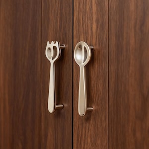 Peut inclure: Gros plan sur deux poignées de porte d'armoire, l'une en forme de fourchette, l'autre de cuillère. Elles sont de couleur argentée et fixées sur une armoire en bois brun foncé. Ces poignées ajoutent une touche unique à la cuisine.