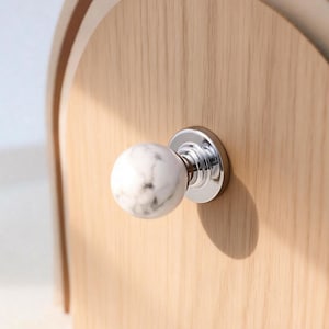 Op de afbeelding: Een houten kast met een marmeren en chromen knop. De knop is een witte bol met grijze adering, gemonteerd op een glanzende chromen basis. De kast is gemaakt van lichtgekleurd hout.