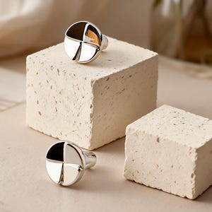 May include: Two silver cufflinks with a segmented design, each divided into four sections of alternating black and silver. The cufflinks are displayed on a textured, off-white surface, with a neutral background.