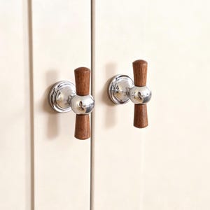 May include: Two cabinet pulls with a polished chrome base and a wooden handle. The handles are a dark brown colour and are cylindrical in shape. The pulls are mounted on a cream-coloured cabinet door.