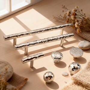 Peut inclure: Une collection de quincaillerie d'armoire argentée, comprenant des barres de tirage, une poignée en T et divers styles de boutons. Le matériel présente une texture martelée, reflétant la lumière. Les articles sont disposés sur une surface en bois de couleur claire.