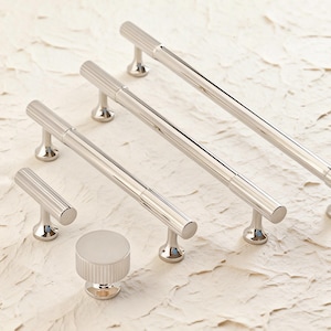 May include: A collection of polished silver cabinet hardware, including several bar pulls and a round knob. The hardware features a ribbed design on the cylindrical portions and is set against a neutral background.