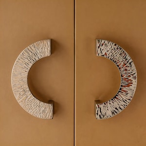 May include: Two silver cabinet handles with a textured, semi-circular design. The handles are mounted on a light brown cabinet door. The handles have a unique, artistic appearance, adding a decorative element to the cabinet.