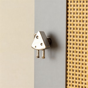 Op de afbeelding: Een messingkleurige kastknop in de vorm van een driehoek, met twee kleine pootjes en drie cirkelvormige inkepingen. Het heeft een lus aan de bovenkant. De knop is gemonteerd op een grijze ondergrond, met rechts een geweven patroon.