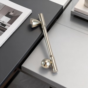 以下が含まれることがあります： 幾何学的なデザインの、磨かれたシルバーのボトルストッパー。ストッパーは円錐形のトップ、円筒形のステム、球形のベースが特徴です。グレーの表面に置かれています。