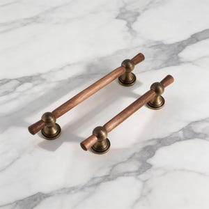 Puede incluir: Dos tiradores de armario de madera con herrajes de color latón. Los tiradores tienen una barra cilíndrica de madera y se fijan al armario con una base redonda y decorativa. Los herrajes tienen un aspecto vintage y antiguo.