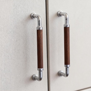 Puede incluir: Dos tiradores de armario con un diseño moderno. Cada tirador presenta un tirador de metal plateado pulido y un agarre cilíndrico de madera marrón oscuro. Los tiradores están montados sobre una superficie clara y texturizada.