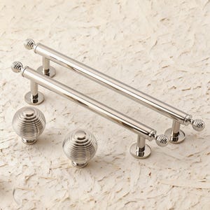 May include: A set of polished nickel cabinet hardware, including two bar pulls and two round knobs. The bar pulls have a cylindrical design with decorative ball ends. The knobs feature a ribbed, spherical design.