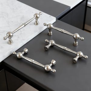 Op de afbeelding: Verschillende gepolijste nikkelen kastgrepen met een klassiek ontwerp. Elke greep heeft een centrale balk met decoratieve balaccenten aan elk uiteinde en op de palen. De grepen zijn gerangschikt op een wit en grijs oppervlak.