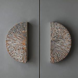 May include: A pair of semi-circular cabinet handles with a textured, sunburst pattern. The handles have a light colour with a distressed, white finish and a warm, brown undertone. They are mounted on a grey cabinet door.